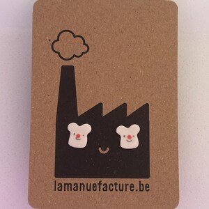 Könnte beinhalten: Weiße Ohrstecker in Bärenform mit roten Nasen auf einer braunen Karte. Die Karte zeigt eine schwarze Fabrikillustration mit einem lächelnden Gesicht und dem Text "lamanufacture.be".
