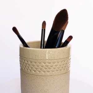 Op de afbeelding: Een beige keramische make-up kwastenhouder met een textuurontwerp. Vier make-up kwasten bevinden zich in de houder.