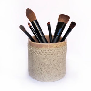 Op de afbeelding: Een set make-upkwasten in een gespikkelde beige keramische houder. De kwasten hebben zwarte handvatten en bruine haren.