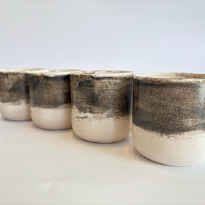 Puede incluir: Cuatro tazas de cerámica con un diseño de dos tonos. La mitad inferior de cada taza es de color blanco crema, mientras que la mitad superior es de color gris oscuro texturizado y moteado. Las tazas están dispuestas en fila, mostrando su diseño simple y moderno.