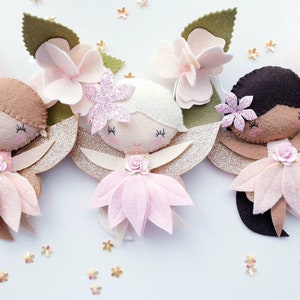 Three felt fairy ornaments with pink flower dresses , gold glitter wings, and flowers. The fairies have different hair colors: brown, white, and black.