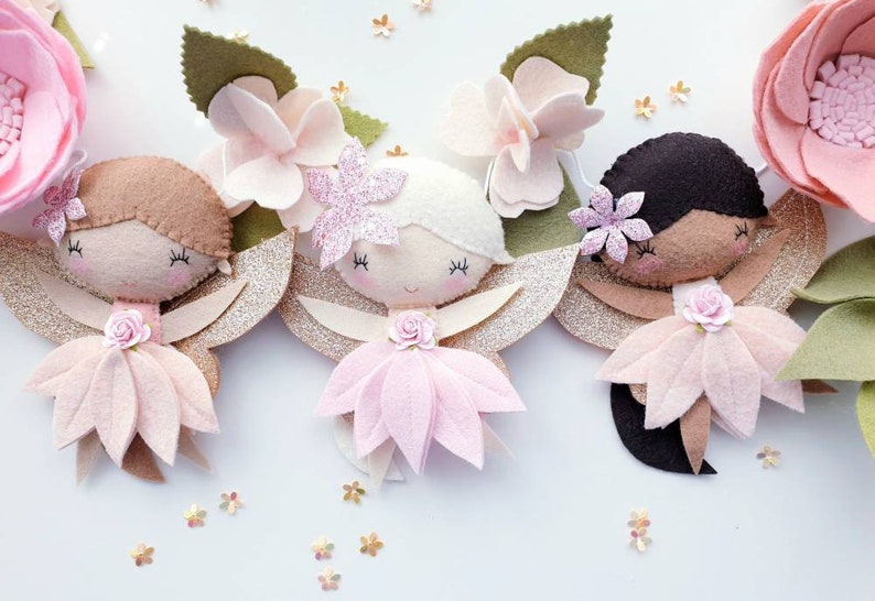 Three felt fairy ornaments with pink flower dresses , gold glitter wings, and flowers. The fairies have different hair colors: brown, white, and black.