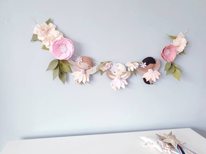 The garland features three pink felt fairies with glitter wings and different colour hair and skin tone. Each has a hand embroidered face and lightly blushed cheeks. On each side of the fairies are large pink felt flowers.
