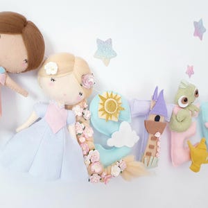 May include: A decorative felt banner featuring two fabric dolls with brown and blonde hair, alongside letters spelling out a name. The banner includes felt icons of a sun, a tower, a green lizard, a gold lamp, and small clouds, accented with pink flowers.