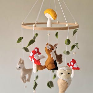 May include: A wooden baby mobile featuring felt woodland creatures, including a fox, owl, deer, and mushrooms. The mobile hangs from a wooden ring and has green felt leaves attached.