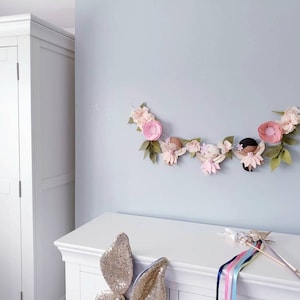 The garland features three pink felt fairies with glitter wings and different colour hair and skin tone. Each has a hand embroidered face and lightly blushed cheeks. On each side of the fairies are large pink felt flowers.