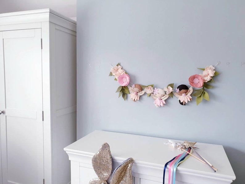 The garland features three pink felt fairies with glitter wings and different colour hair and skin tone. Each has a hand embroidered face and lightly blushed cheeks. On each side of the fairies are large pink felt flowers.