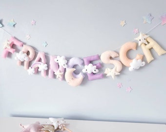 May include: A pastel-colored felt banner with the name "FRANCESCA" spelled out in individual letters. Each letter is decorated with a small cloud, star, or crescent moon.