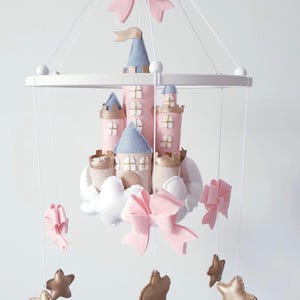 May include: A pink and blue felt castle mobile with white clouds, gold stars, and pink bows. The castle has multiple towers and a flag. The mobile hangs from a white wooden frame with white beads.