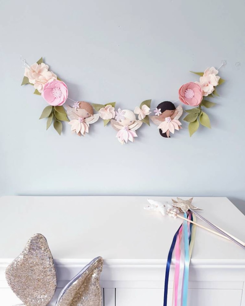 The garland features three pink felt fairies with glitter wings and different colour hair and skin tone. Each has a hand embroidered face and lightly blushed cheeks. On each side of the fairies are large pink felt flowers.