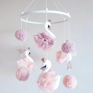 Pode incluir: Um móbile de bebê branco com detalhes em rosa e dourado. O móbile apresenta três cisnes brancos com saias de tule rosa e coroas douradas, pompons rosas e estrelas douradas com glitter.