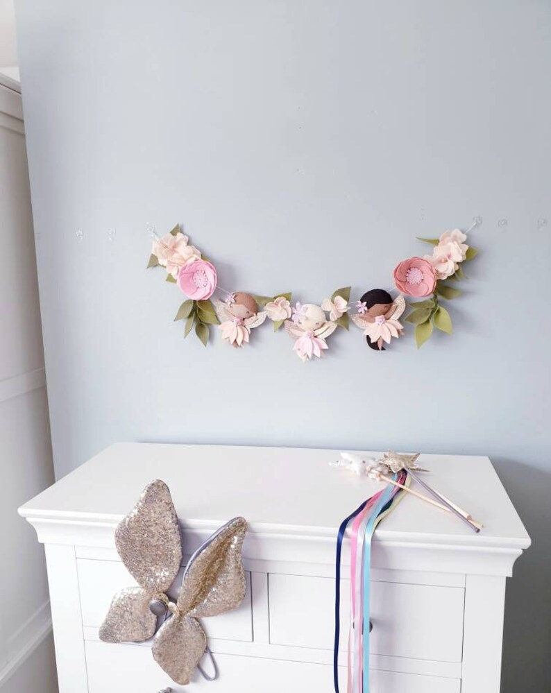 The garland features three pink felt fairies with glitter wings and different colour hair and skin tone. Each has a hand embroidered face and lightly blushed cheeks. On each side of the fairies are large pink felt flowers.