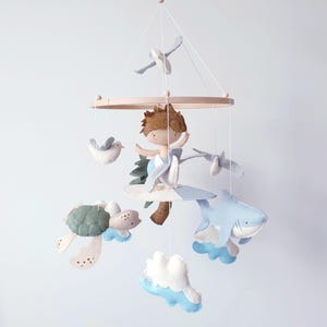 May include: A wooden baby mobile featuring a surfer boy, a shark, a turtle, clouds, and birds. The mobile is crafted from felt and is in blue, green, brown, and white colours.