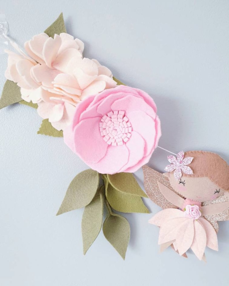 A felt garland with a pink flower, green leaves, and a pink fairy with glitter wings. The fairy has brown hair, a pink flower on her head, and a pink dress.