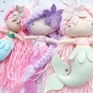 Può includere: Tre bambole sirena fatte a mano con capelli di filo rosa, viola e rosa chiaro. Ogni bambola ha una coda di feltro blu, rosa e verde e un copricapo glitterato. Le bambole hanno tratti del viso ricamati e sono disposte su uno sfondo bianco.