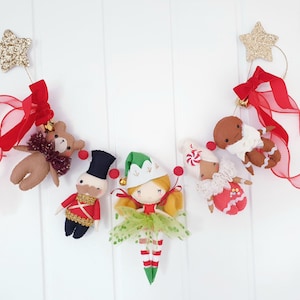 Pode incluir: Uma guirlanda de Natal festiva com enfeites de feltro. A guirlanda inclui um ursinho de pelúcia, um soldado de brinquedo, um elfo e bonecos de gengibre. Cada enfeite é suspenso por uma fita vermelha e uma estrela dourada. Os enfeites são em tons de marrom, vermelho, verde e branco.