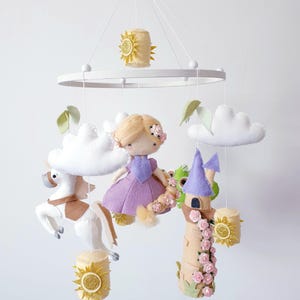 May include: A handmade baby mobile with a princess doll in a purple dress, a white horse, a castle, and clouds. The mobile includes gold sun accents and is suspended from a white circular frame. The design is soft and whimsical.