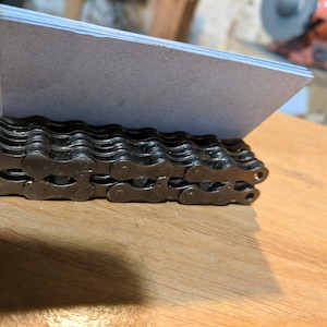 May include: A black metal business card holder made from a bicycle chain. The holder is sitting on a wooden surface.