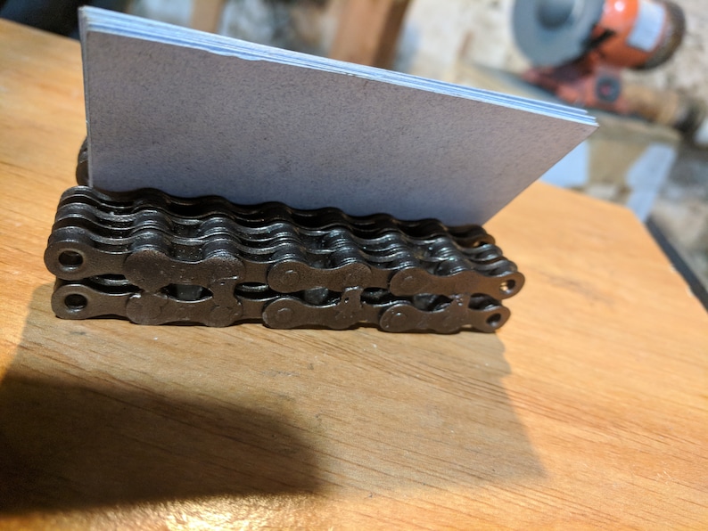 May include: A black metal business card holder made from a bicycle chain. The holder is sitting on a wooden surface.