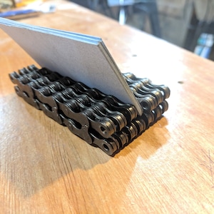 May include: A black metal business card holder made from stacked bicycle chains. The holder is sitting on a wooden surface and has a grey card inside.