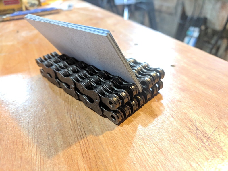 May include: A black metal business card holder made from stacked bicycle chains. The holder is sitting on a wooden surface and has a grey card inside.