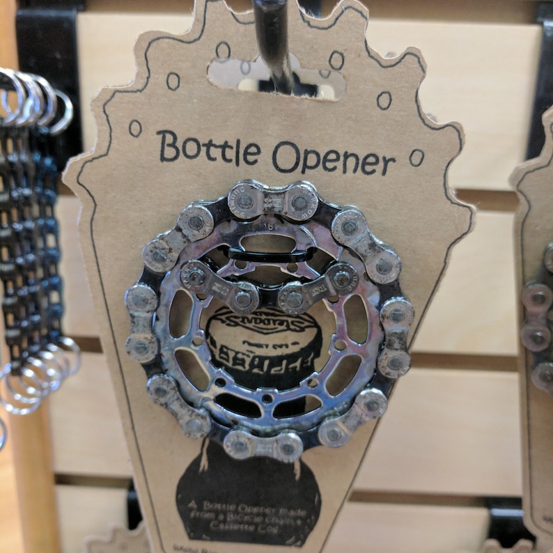 May include: A silver bottle opener made from a bicycle chain cassette. The opener is attached to a brown cardboard tag with the text "Bottle Opener" printed on it.