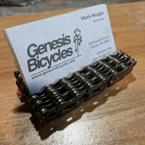 May include: A black metal business card holder made from a bicycle chain. The holder is holding a business card for Genesis Bicycles, a bicycle shop in Easton, Pennsylvania. The business card includes the shop's website, phone number, and fax number.