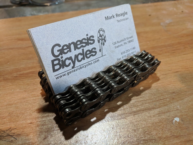 May include: A black metal business card holder made from a bicycle chain. The holder is holding a business card for Genesis Bicycles, a bicycle shop in Easton, Pennsylvania. The business card includes the shop's website, phone number, and fax number.