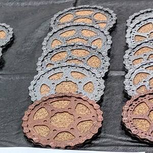 May include: A set of eight round coasters made from cork and metal. The coasters are stacked in two piles, one with silver metal and one with copper metal. The coasters have a circular design with a gear-like pattern.