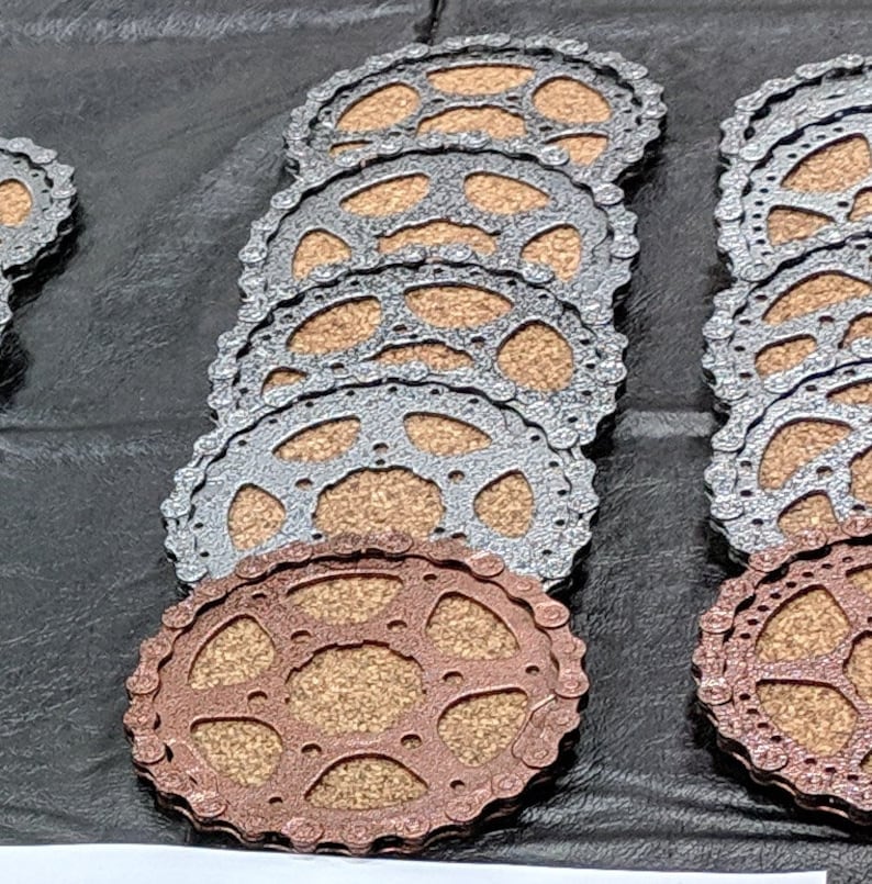 May include: A set of eight round coasters made from cork and metal. The coasters are stacked in two piles, one with silver metal and one with copper metal. The coasters have a circular design with a gear-like pattern.