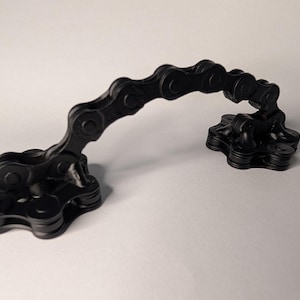 May include: A matte black handle constructed from interconnected metal bicycle chain links. The handle features two circular bases, each composed of layered chain segments, connected by an arched section of chain that creates a curved grip.