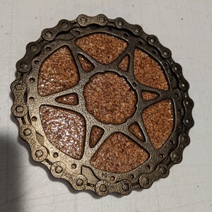 May include: A round cork coaster with a metal design of a bicycle chain and a starburst pattern. The coaster is brown and gold.