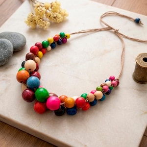 May include: A colorful wooden bead necklace with a variety of hues, including red, green, blue, and pink. The necklace is strung on a natural cord and is displayed on a marble surface. A decorative spool and stones are also present.