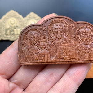 May include: A brown, rectangular, arched-top religious plaque featuring a relief carving of three figures, including a central figure holding a book, a figure with a child, and a figure wearing a crown, with Greek lettering inscribed throughout.