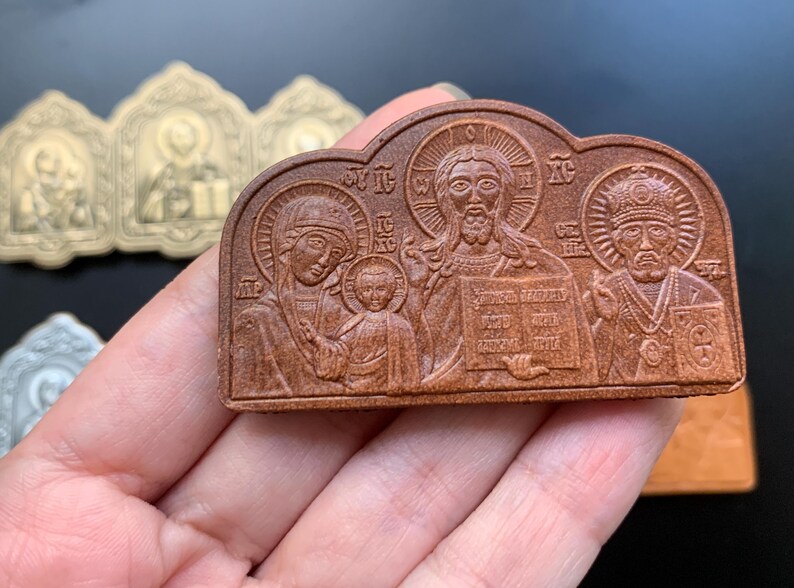May include: A brown, rectangular, arched-top religious plaque featuring a relief carving of three figures, including a central figure holding a book, a figure with a child, and a figure wearing a crown, with Greek lettering inscribed throughout.
