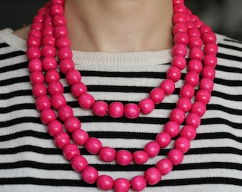 May include: A layered necklace featuring three strands of large, round, hot pink wooden beads. The necklace is displayed against a black and white striped shirt.
