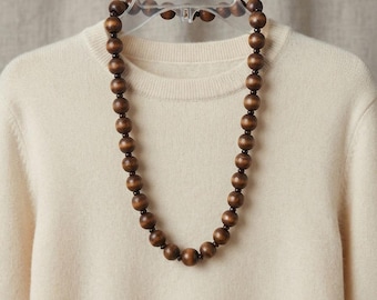 May include: A long necklace featuring large, round, dark brown wooden beads strung together with smaller, dark brown spacer beads. The necklace is displayed on a cream-colored, long-sleeved knit sweater hanging on a clear plastic hanger.