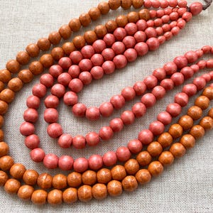 Può includere: Una collezione di collane di perline di legno nei toni dell'arancione e del corallo. Le perline rotonde sono infilate, creando un effetto a strati. Le collane sono esposte su una superficie di colore neutro.