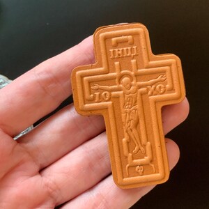 May include: A tan-colored, cross-shaped object featuring an embossed depiction of a figure with arms outstretched. The design includes the Greek letters IC and XC on either side of the figure, along with additional text at the top and bottom.