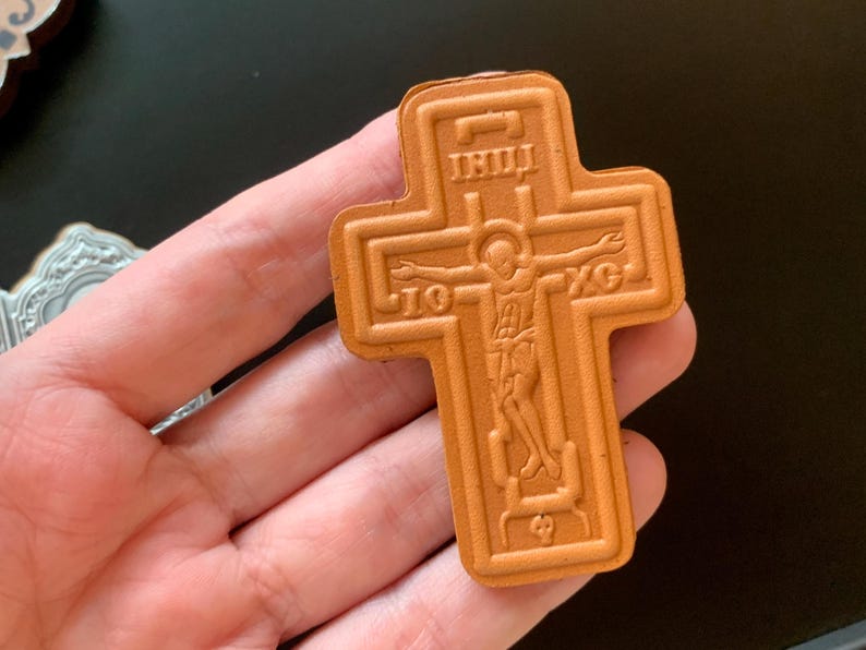May include: A tan-colored, cross-shaped object featuring an embossed depiction of a figure with arms outstretched. The design includes the Greek letters IC and XC on either side of the figure, along with additional text at the top and bottom.