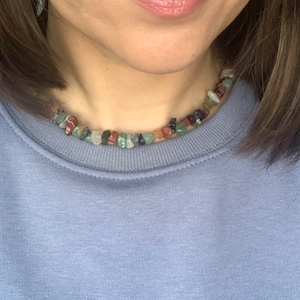 K&ouml;nnte beinhalten: Nahaufnahme einer Frau, die ein hellblaues Sweatshirt mit einer mehrfarbigen Stein-Halskette tr&auml;gt. Die Halskette besteht aus kleinen, unregelm&auml;&szlig;ig geformten Steinen in Gr&uuml;n-, Braun-, Rot- und Schwarzt&ouml;nen.