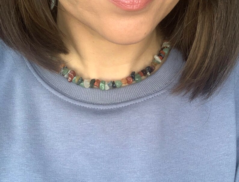 K&ouml;nnte beinhalten: Nahaufnahme einer Frau, die ein hellblaues Sweatshirt mit einer mehrfarbigen Stein-Halskette tr&auml;gt. Die Halskette besteht aus kleinen, unregelm&auml;&szlig;ig geformten Steinen in Gr&uuml;n-, Braun-, Rot- und Schwarzt&ouml;nen.