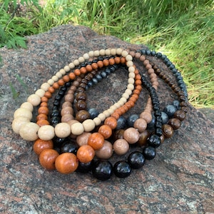 May include: A collection of multiple beaded necklaces featuring spherical beads in various shades of light beige, medium brown, dark brown, black, and grey. The beads are arranged in graduated sizes, with larger beads at the center and smaller beads toward the ends.