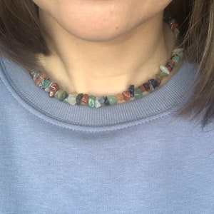 K&ouml;nnte beinhalten: Nahaufnahme einer Person, die ein hellblaues Sweatshirt mit einer mehrfarbigen Stein-Halskette tr&auml;gt. Die Halskette besteht aus kleinen, unregelm&auml;&szlig;ig geformten Steinen in Rot-, Gr&uuml;n-, Braun- und Schwarzt&ouml;nen.