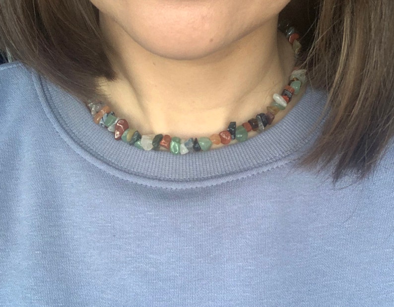 K&ouml;nnte beinhalten: Nahaufnahme einer Person, die ein hellblaues Sweatshirt mit einer mehrfarbigen Stein-Halskette tr&auml;gt. Die Halskette besteht aus kleinen, unregelm&auml;&szlig;ig geformten Steinen in Rot-, Gr&uuml;n-, Braun- und Schwarzt&ouml;nen.