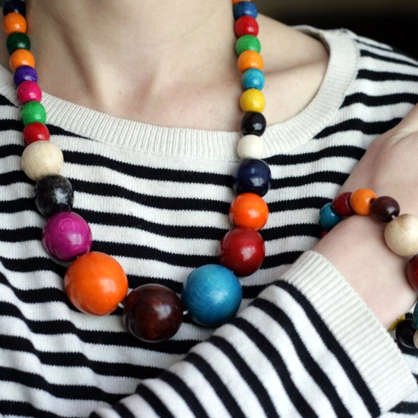 May include: A colorful wooden bead necklace and bracelet set. The necklace is long and features large, round beads in various colors, including red, orange, yellow, green, blue, purple, and black. The bracelet is shorter and features smaller, round beads in similar colors.