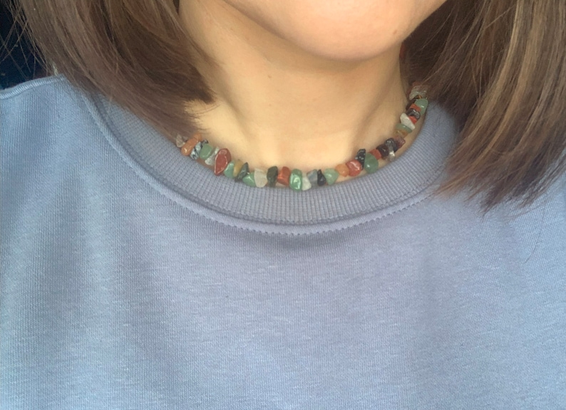K&ouml;nnte beinhalten: Nahaufnahme einer Person, die ein hellblaues Sweatshirt mit einer mehrfarbigen Stein-Halskette tr&auml;gt. Die Halskette besteht aus kleinen, unregelm&auml;&szlig;ig geformten Steinen in Gr&uuml;n-, Rot-, Braun- und Schwarzt&ouml;nen.