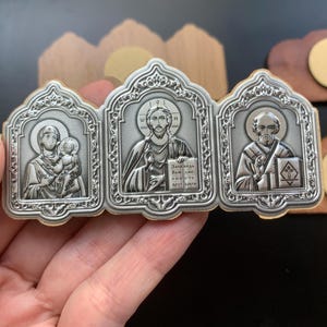 May include: A triptych icon featuring three silver-toned metal panels with embossed religious figures. The left panel shows a figure holding a child, the center shows a central figure holding a book with Cyrillic text, and the right shows a bearded figure.