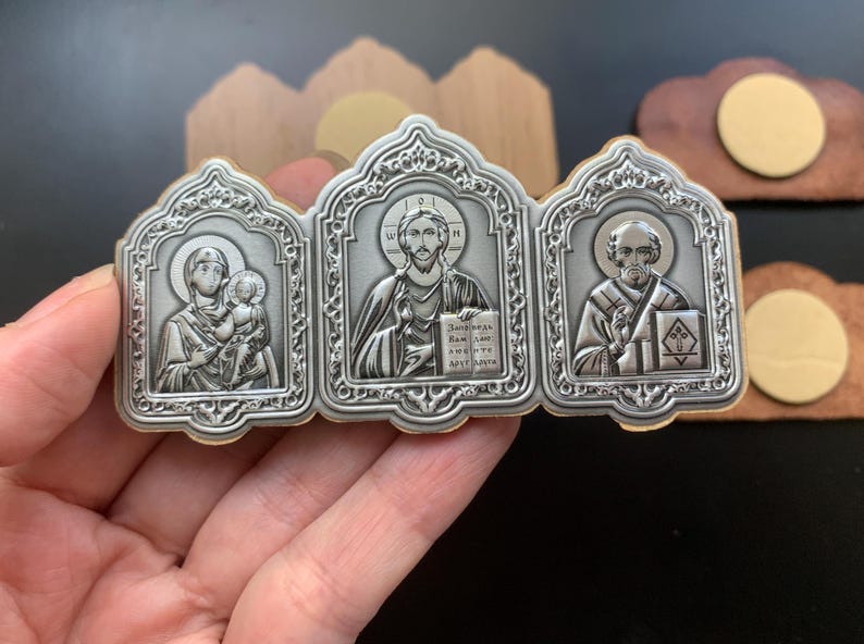May include: A triptych icon featuring three silver-toned metal panels with embossed religious figures. The left panel shows a figure holding a child, the center shows a central figure holding a book with Cyrillic text, and the right shows a bearded figure.