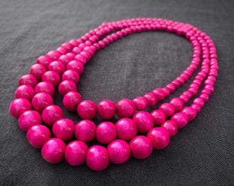 May include: A multi-strand necklace featuring three layers of round, vibrant hot pink wooden beads. The beads are uniform in shape and size, strung together to create a layered effect with a smooth, polished finish on each individual bead.